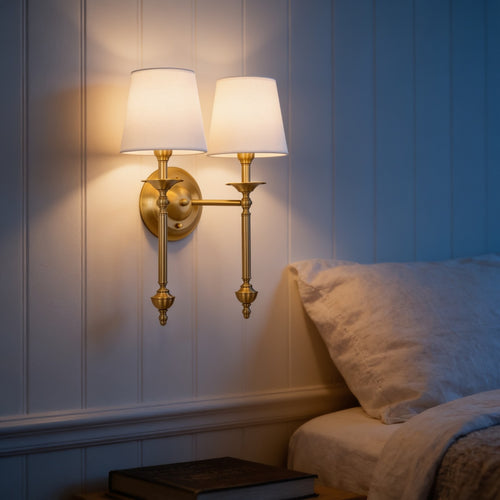 Solid brass two-arm wall sconce with white fabric shades beside a bed
